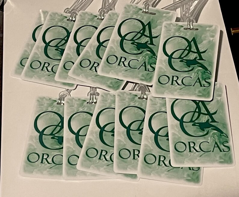 Może przedstawiać: Kolekcja prostokątnych etykiet bagażowych z zielono-białym marmurkowym wzorem. Każda etykieta zawiera napis "ORCAS" oraz grafikę orki w ciemnozielonym kolorze. Etykiety posiadają przezroczyste plastikowe pętle do mocowania.