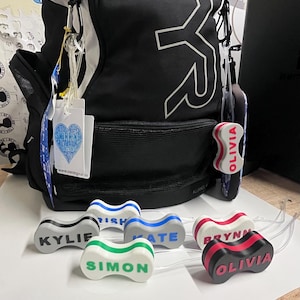 May include: A black TYR backpack with white lettering and several personalized swim goggles. The goggles are in various colors, including gray, blue, green, and red, each with a name printed on them. A tag with a heart-shaped logo is attached.