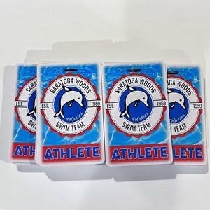Może przedstawiać: Cztery prostokątne odznaki sportowc&oacute;w. Każda odznaka zawiera logo delfina w okrągłym emblemacie z napisem "SARATOGA WOODS SWIM TEAM" i "1959". Słowo "ATHLETE" jest wydrukowane pogrubionymi czerwonymi literami na dole każdej odznaki.