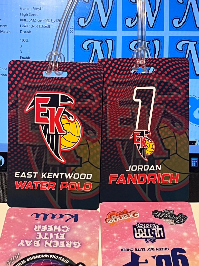 Może przedstawiać: Dwie czerwono-czarne identyfikatory drużyny piłki wodnej z napisem "EAST KENTWOOD WATER POLO" i "JORDAN FANDRICH". Identyfikatory mają czerwono-czarne tło z motywem koszyk&oacute;wki. Poniżej znajdują się dwa r&oacute;żowe identyfikatory cheerleaderek.
