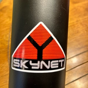 May include: Black water bottle with a white sticker featuring a red triangle with a black Y inside and the text "SKYNET" underneath.