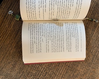 以下が含まれることがあります： 緑と茶色のチャームが付いたしおりが付いた開いた本のクローズアップ。本は、キャプテンとエディというキャラクターについて書かれたページを開いています。テキストには、「なぜなら、彼は落ち着いて、エディの胸に肘を置きながら言った、「私たちはあなたをその火事で失っていたでしょう。あなたは死んでいたでしょう。そして、それはあなたの時間ではありませんでした。」と書いてあります。