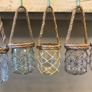 Può includere: Sei barattoli di vetro con manici in corda e un design a rete metallica. I barattoli sono appesi a una superficie di legno bianco. I barattoli sono di diversi colori, tra cui blu, verde, giallo e viola.