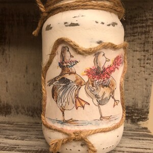 Può includere: Un barattolo di vetro dipinto di bianco con un design in corda rustico. Il barattolo presenta un'immagine dipinta a mano di due anatre con accenti colorati.