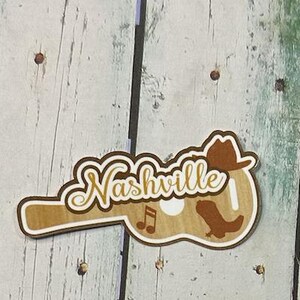 May include: A brown wooden guitar-shaped sign with the word "Nashville" in gold lettering. The sign also features a brown cowboy hat, a brown boot, and musical notes.