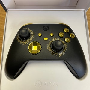 Może przedstawiać: Czarny kontroler Xbox z złotymi akcentami i okrągłym wzorem z elficką czcionką. Kontroler znajduje się w oryginalnym opakowaniu.