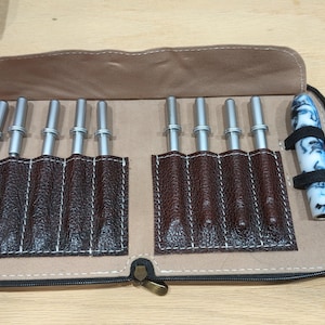 May include: A brown leatherette case with a zipper, holding multiple silver tools with brown leather pockets. A pen with a blue and white swirl design is secured with a black strap. The case is open on a light brown surface.