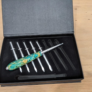 May include: A set of crochet hooks in a black box. The hooks have silver metal shafts and colorful, patterned handles in shades of teal, yellow, and green. The box is open, revealing the hooks arranged neatly.