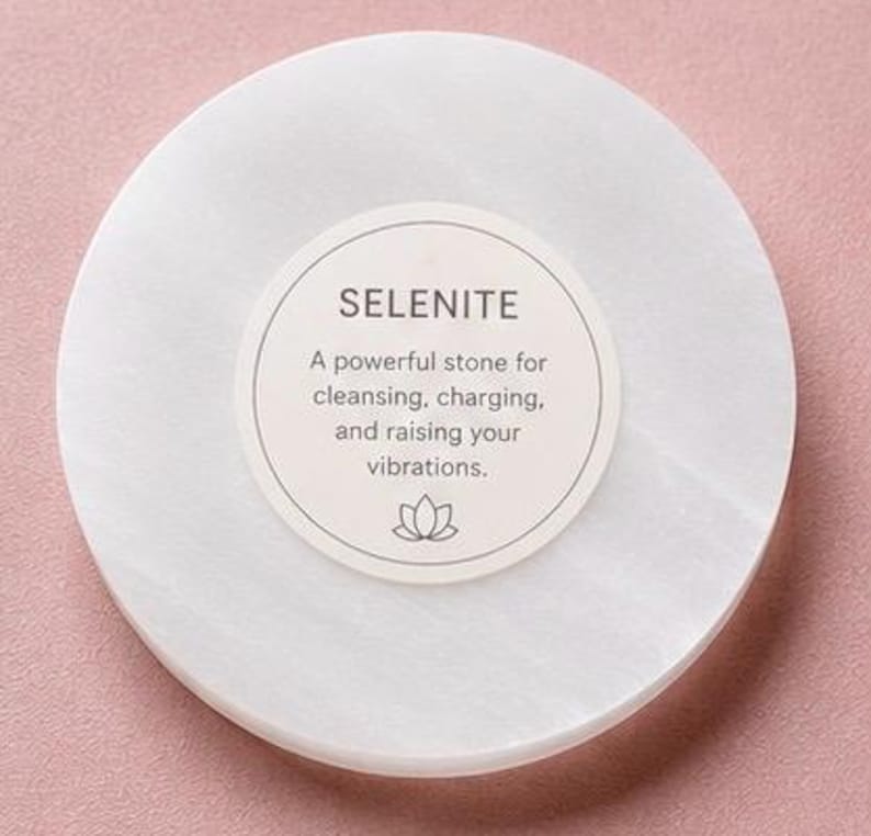 May include: A circular white selenite stone slab featuring a centered circular label with the text SELENITE, A powerful stone for cleansing, charging, and raising your vibrations, and a small lotus flower icon at the bottom of the label.