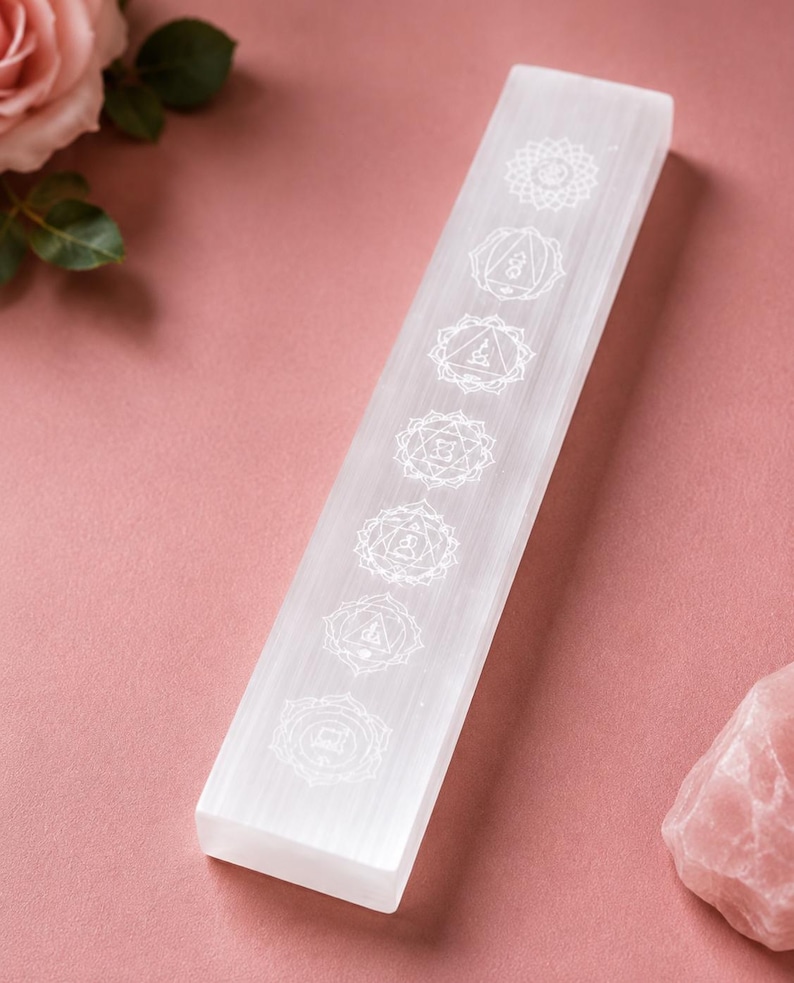 May include: A rectangular selenite crystal wand featuring seven white engraved chakra symbols arranged in a vertical line along its smooth, translucent surface. The crystal has a clear, fibrous texture and a flat, elongated shape.