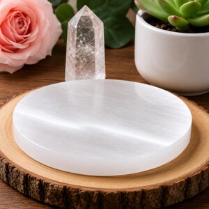 May include: A circular, white selenite charging plate with a smooth, fibrous texture. The disc is positioned flat, showcasing its reflective, pearlescent surface and clean, rounded edges, providing a neutral base for displaying various small objects.