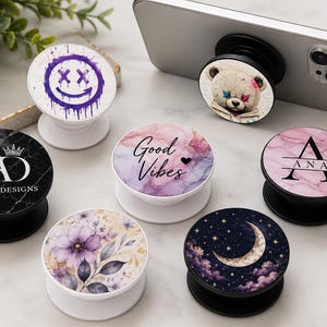 May include: A collection of six circular phone grips featuring various designs including a purple dripping smiley face, a teddy bear, a black marble pattern with the text ANA'S DESIGNS, a pink marble pattern with the name ANA, a floral print, and a moon.
