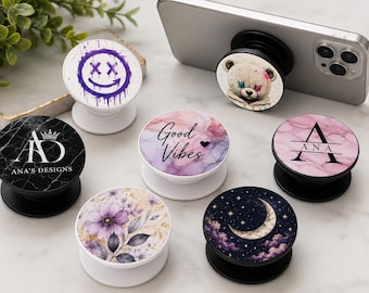 May include: A collection of six circular phone grips featuring various designs including a purple dripping smiley face, a teddy bear, a black marble pattern with the text ANA'S DESIGNS, a pink marble pattern with the name ANA, a floral print, and a moon.