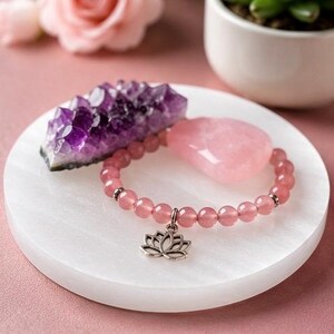 May include: A beaded bracelet made of translucent pink round stones featuring a silver-toned lotus flower charm. The bracelet is displayed alongside a jagged purple amethyst crystal cluster and a smooth, heart-shaped pink rose quartz stone.