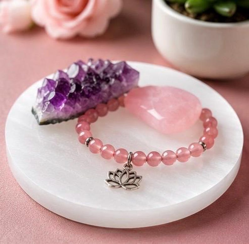 May include: A beaded bracelet made of translucent pink round stones featuring a silver-toned lotus flower charm. The bracelet is displayed alongside a jagged purple amethyst crystal cluster and a smooth, heart-shaped pink rose quartz stone.