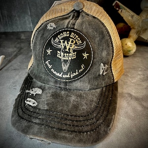 May include: Distressed black and tan trucker hat featuring a patch with the text "WRONG BITCH RANCH" and a bull skull graphic. The hat has a mesh back and a curved brim with distressed details.