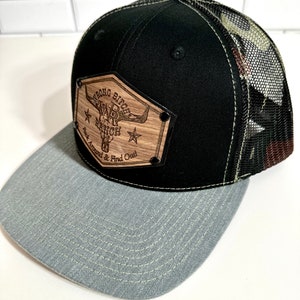 Puede incluir: Una gorra de camionero de camuflaje negro y gris con un parche de madera que dice "Strong Bitch Ranch, Buck Around & Find Out".