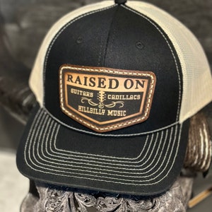 May include: Black and white trucker hat with a brown leather patch that says "Raised On Guitars & Cadillacs Hillbilly Music".