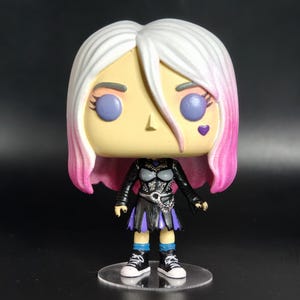 Op de afbeelding: Een gestileerde vinyl figuur met een groot hoofd en een klein lichaam. De figuur heeft wit haar met roze punten, paarse ogen en een hartvormig teken. Het draagt een zwarte outfit met een rok, ketting en sneakers, staand op een heldere basis.