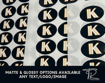 Könnte beinhalten: Ein Blatt weißes Papier mit mehreren schwarzen und goldenen ovalen Aufklebern. Jeder Aufkleber zeigt einen goldenen Buchstaben "K" auf schwarzem Hintergrund. Der Text "MATTE & GLOSSY OPTIONS AVAILABLE ANY TEXT/LOGO/IMAGE" ist auf das Papier gedruckt.