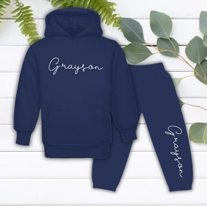 May include: Navy blue hooded sweatshirt and sweatpants set with the name "Grayson" written in white cursive on the front of the hoodie and the leg of the sweatpants.