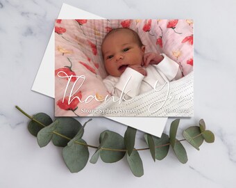 May include: Thank You card with a photo of a newborn baby girl in a pink and white floral swaddle. The card says "Thank You" in white script with the baby's name "Sloane Sydney Symons" and the date "08.01.2021" below.