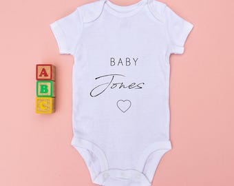 May include: A white short-sleeved baby bodysuit featuring the text BABY Jones in black lettering with a small heart outline below. Beside the garment is a stack of three small wooden blocks with the letters A, B, and C printed on them.