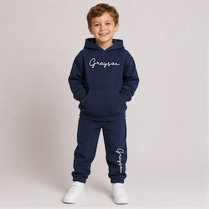 May include: A navy blue hooded sweatshirt and matching sweatpants set. The hoodie features the name Grayson in white cursive script on the chest. The sweatpants feature the same name Grayson in white cursive script on the lower left leg.