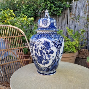 Könnte beinhalten: Eine große blaue und weiße Keramikvase mit Deckel und aufwendigen Blumendesigns. Die Vase steht auf einem Tisch mit einer gewebten Oberfläche.