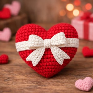 Puede incluir: Un corazón rojo tejido a ganchillo, adornado con un lazo y una banda blancos. Otros pequeños corazones tejidos a ganchillo rojos y rosas están dispersos. El fondo es una superficie de madera borrosa, evocando un ambiente romántico.