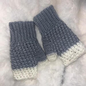 Peut inclure: Une paire de mitaines, crochetées à la main avec un point texturé. Les gants sont principalement de couleur gris clair, avec un poignet blanc contrastant. Les gants sont présentés sur une surface douce et blanche.