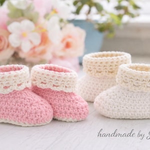 Peut inclure: Bottes de bébé faites à la main en crochet rose et crème. Deux paires de bottines sont présentées, l'une en rose et l'autre en crème. Les bottines ont un design à revers. Les mots "handmade by Julia" sont écrits en cursive.