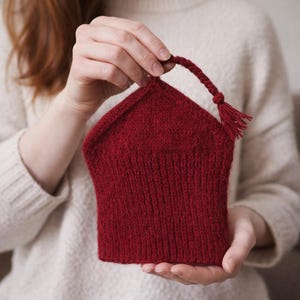 Peut inclure: Un chauffe-thé rouge tricoté, avec une poignée tressée et une gland. Le chauffe-thé est tenu dans les mains d'une personne, mettant en valeur le tricot texturé et la couleur riche et chaleureuse. Conçu pour une théière.