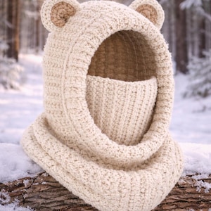 Peut inclure: Cagoule d'ours en crochet de couleur crème avec des oreilles attachées. La cagoule comprend un couvre-visage et une extension en forme d'écharpe. L'article est présenté sur une bûche enneigée dans un cadre forestier hivernal.