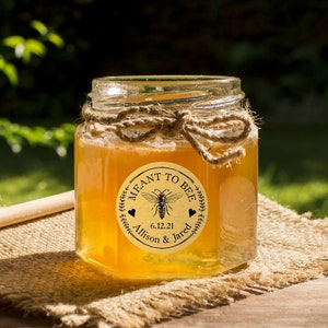 Puede incluir: Un tarro de vidrio de miel con una etiqueta redonda que dice "Meant to Bee" con una ilustración de abeja y la fecha "6.12.21" y los nombres "Alison & Jared".