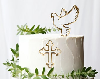 May include: White cake with a gold cross and a gold dove on top. The cake is decorated with green leaves.