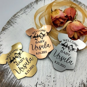 Op de afbeelding: Drie babyshower-cadeautjes in de vorm van een jurk met de tekst "Welkom Baby Ursula 8.21.2021". De cadeautjes zijn gemaakt van goud, zilver en ros&eacute; goud.
