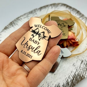 Op de afbeelding: Een koperkleurige houten baby-romper uitsnijding met de tekst "Welcome Baby Ursula 8.21.2021" erop gegraveerd. De romper heeft een strik aan de voorkant.