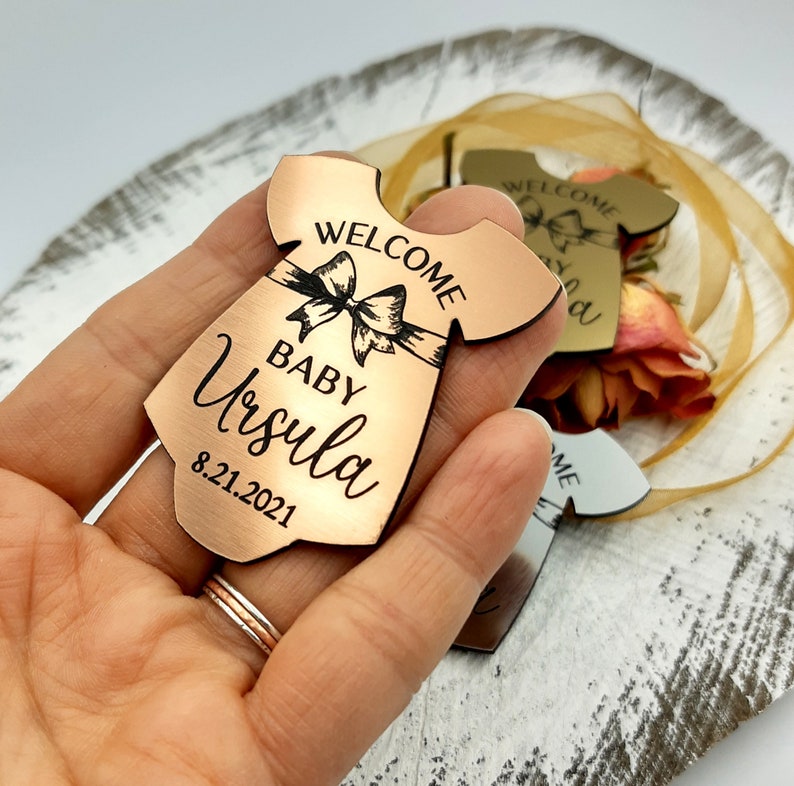 Op de afbeelding: Een koperkleurige houten baby-romper uitsnijding met de tekst "Welcome Baby Ursula 8.21.2021" erop gegraveerd. De romper heeft een strik aan de voorkant.