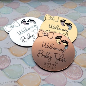 Op de afbeelding: Drie ronde, metalen babyshower-cadeautjes met een strik en babyvoetjes. De cadeautjes zijn zilver, goud en koper. De tekst op de cadeautjes luidt "Welkom Baby Tyler 4.22.23".
