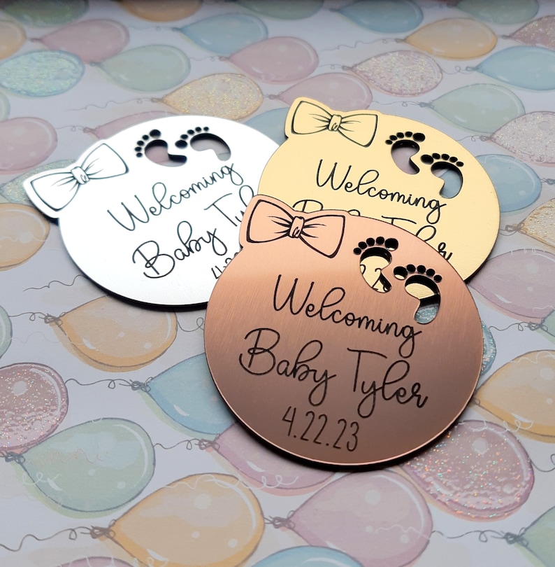 Op de afbeelding: Drie ronde, metalen babyshower-cadeautjes met een strik en babyvoetjes. De cadeautjes zijn zilver, goud en koper. De tekst op de cadeautjes luidt "Welkom Baby Tyler 4.22.23".