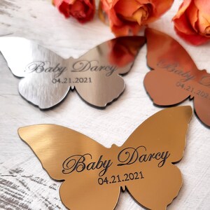 May include: Three silver and gold butterfly-shaped metal ornaments with the text "Baby Darcy 04.21.2021" engraved on each.