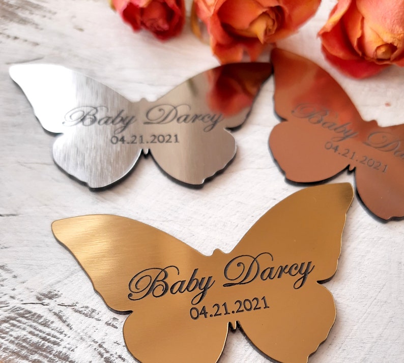 May include: Three silver and gold butterfly-shaped metal ornaments with the text "Baby Darcy 04.21.2021" engraved on each.