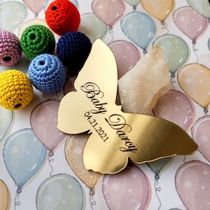 May include: Gold butterfly-shaped keepsake with the text "Baby Darcy 04.21.2021" engraved on it. The keepsake is surrounded by colorful crocheted balls and a white background with a pattern of balloons.