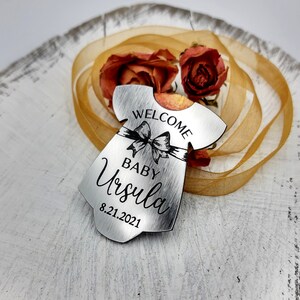 Op de afbeelding: Zilveren metalen babymeisjes ornament in de vorm van een jurk met de tekst "Welcome Baby Ursula 8.21.2021" en een zwarte strik.