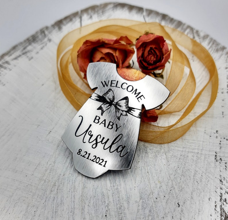Op de afbeelding: Zilveren metalen babymeisjes ornament in de vorm van een jurk met de tekst "Welcome Baby Ursula 8.21.2021" en een zwarte strik.