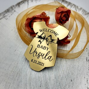 Op de afbeelding: Gouden houten babymeisjesornament in de vorm van een jurk met een zwart lint en strik. Het ornament zegt "Welkom Baby Ursula 8.21.2021".