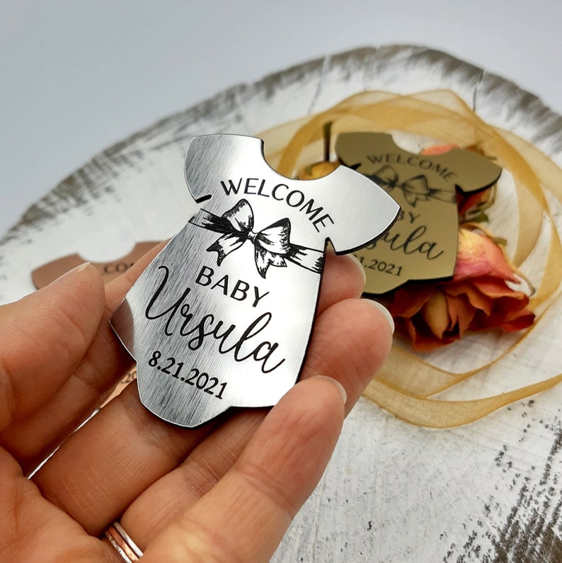 Op de afbeelding: Zilveren metalen baby-rompertje ornament met de tekst "Welkom Baby Ursula 8.21.2021".