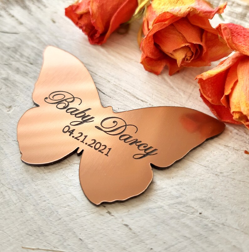 May include: A copper-colored butterfly-shaped metal ornament with the text "Baby Darcy 04.21.2021" engraved on it.