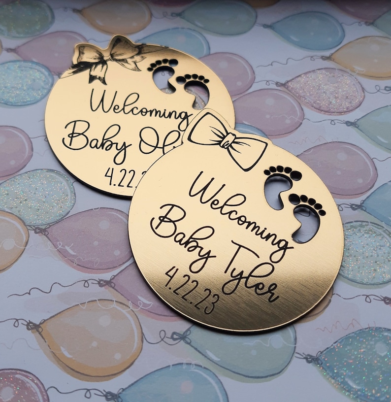 Op de afbeelding: Twee ronde gouden baby-aankondigingsplaquettes met een strik en voetafdrukken. De bovenste plaquette zegt "Welkom Baby Olivia 4.22.23" en de onderste plaquette zegt "Welkom Baby Tyler 4.22.23".